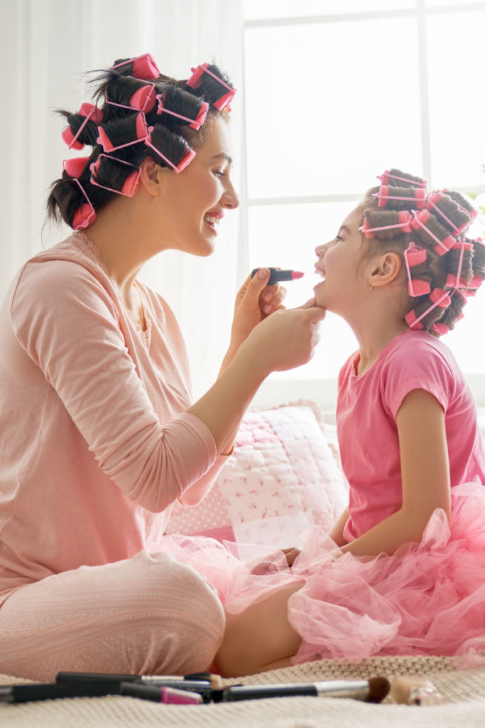 mom and daughter wearing pink with pink rollers in their hair sitting on a bed and mom putting lipstick on the little girl