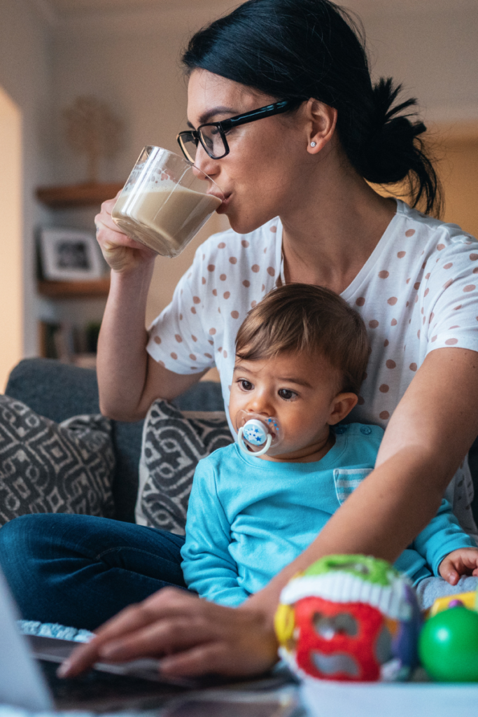 woman drinking coffee, working on a laptop, and holding a baby