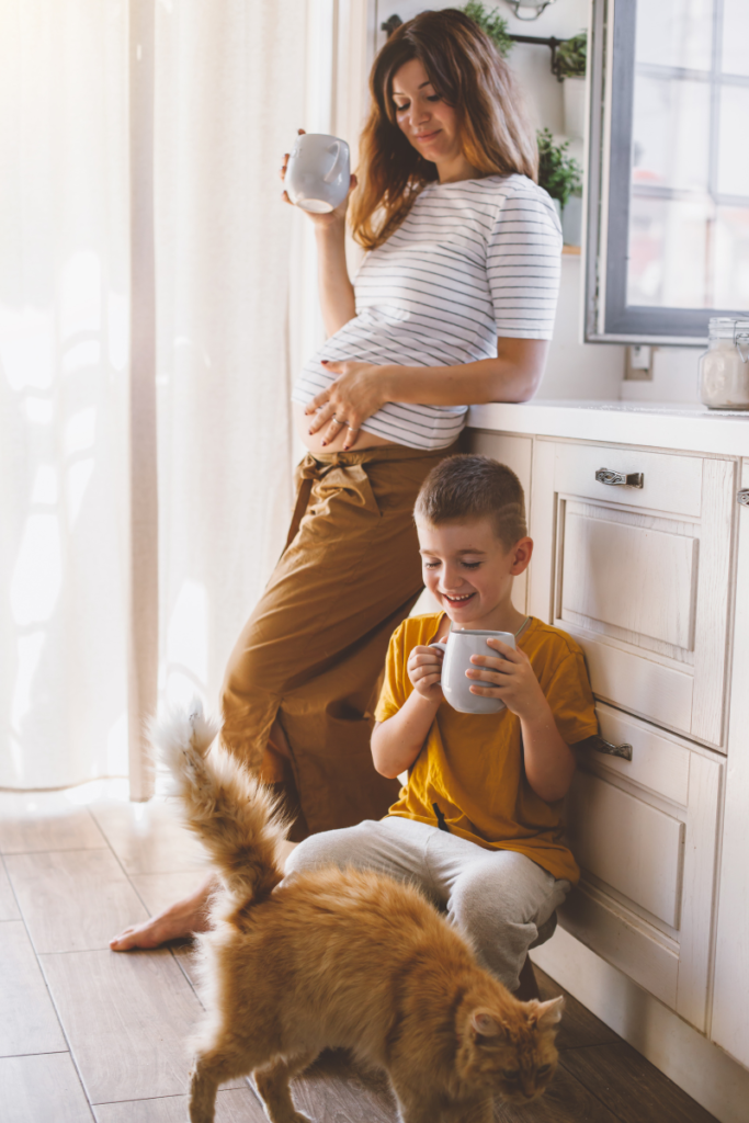 mom and son in the kitchen both holding mugs and looking at an orange cat 