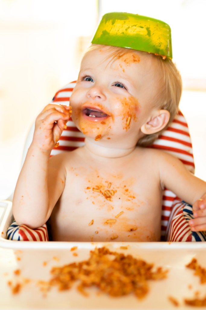 Baby in a high chair covered in spaghetti with a bowl on his head
