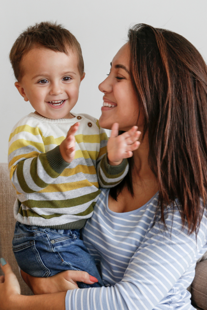 Woman holding a little boy and smiling at him while he looks at the camera and is about to clap
